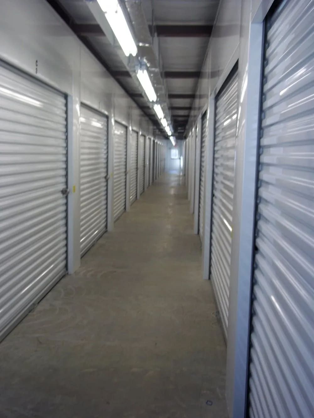 Storage unit hallway with white doors, fluorescent lights, and concrete floor.