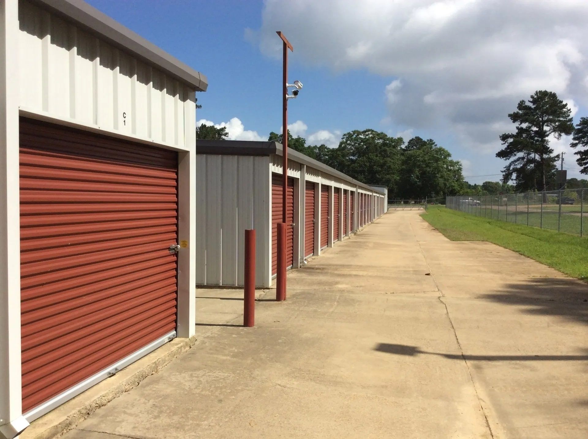 Storage units with red doors, long concrete walkway, surveillance camera, cloudy sky.