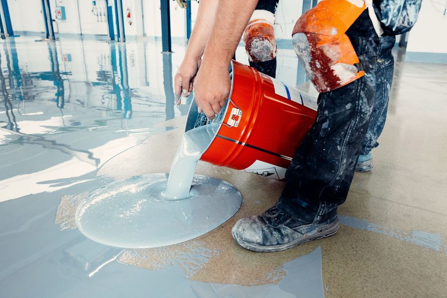 Person pouring light gray epoxy flooring from a red bucket onto a gray floor.