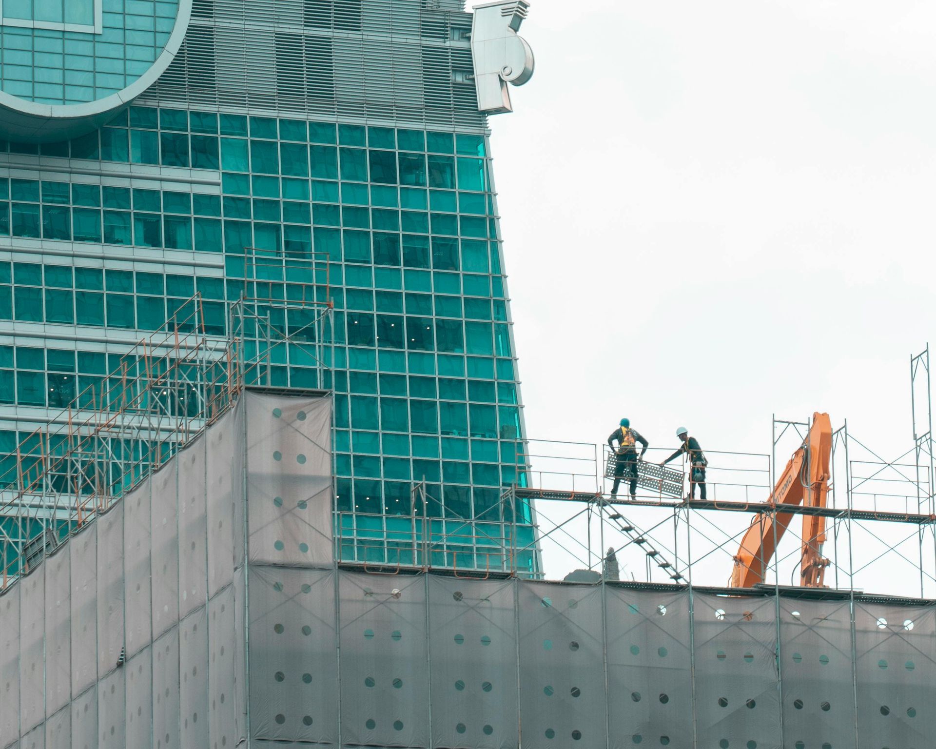 Construction workers on scaffolding at a building site with a tall, teal-glass skyscraper in the background.