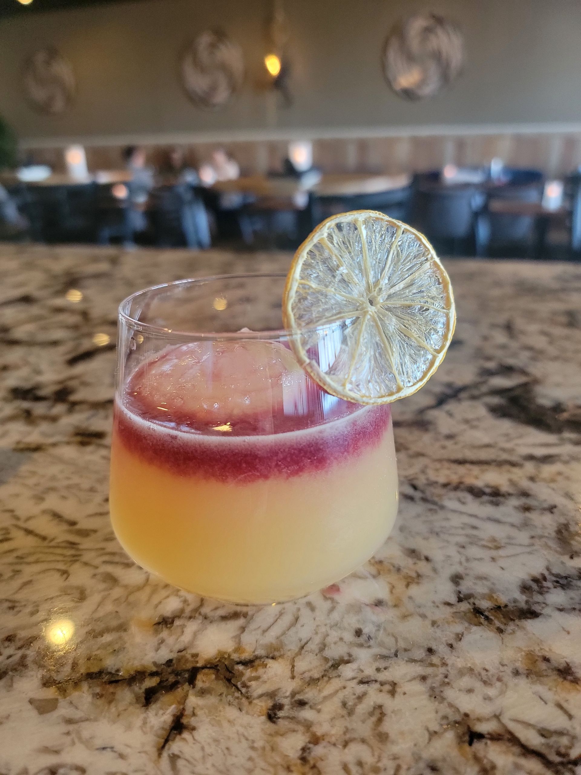 Cocktail in a glass with yellow and purple layers, garnished with a dried citrus slice. Set on a bar.