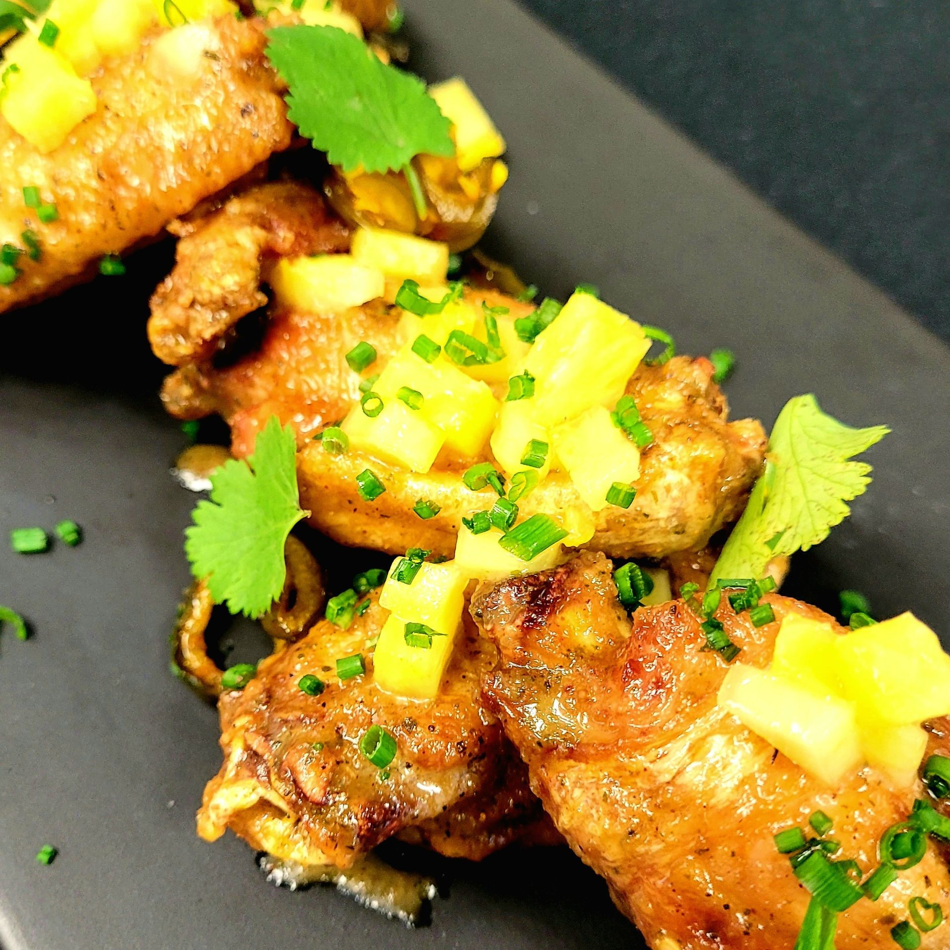 Fried chicken wings topped with pineapple salsa and chives on a black plate.