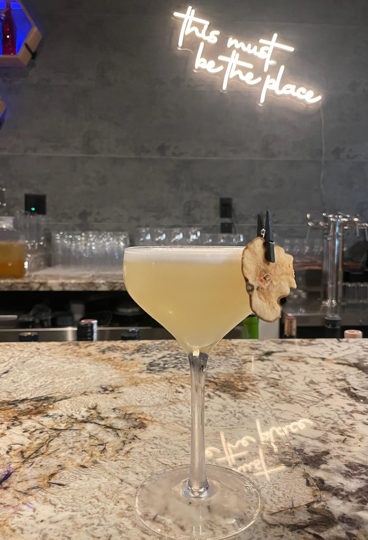 Cocktail in a stemmed glass garnished with dried fruit on a bar with a neon sign, “This must be the place.”
