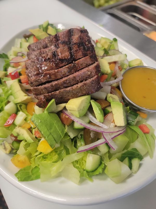 Grilled steak slices atop a salad with avocado, cucumber, red onion, and dressing in a small bowl.