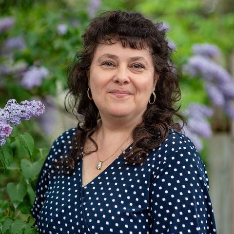 A woman in a blue polka dot shirt is standing in front of purple flowers.