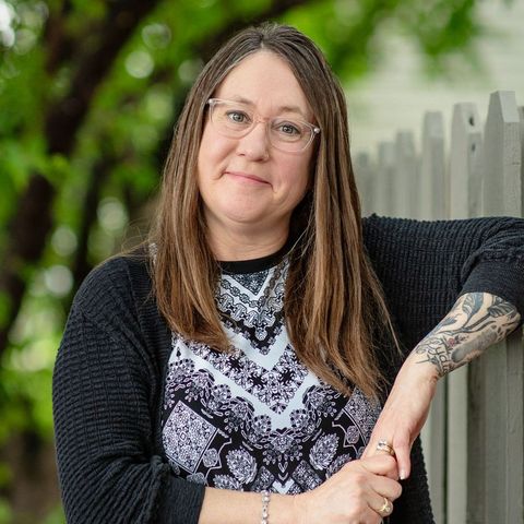 A woman with glasses and a tattoo on her arm is leaning against a fence.