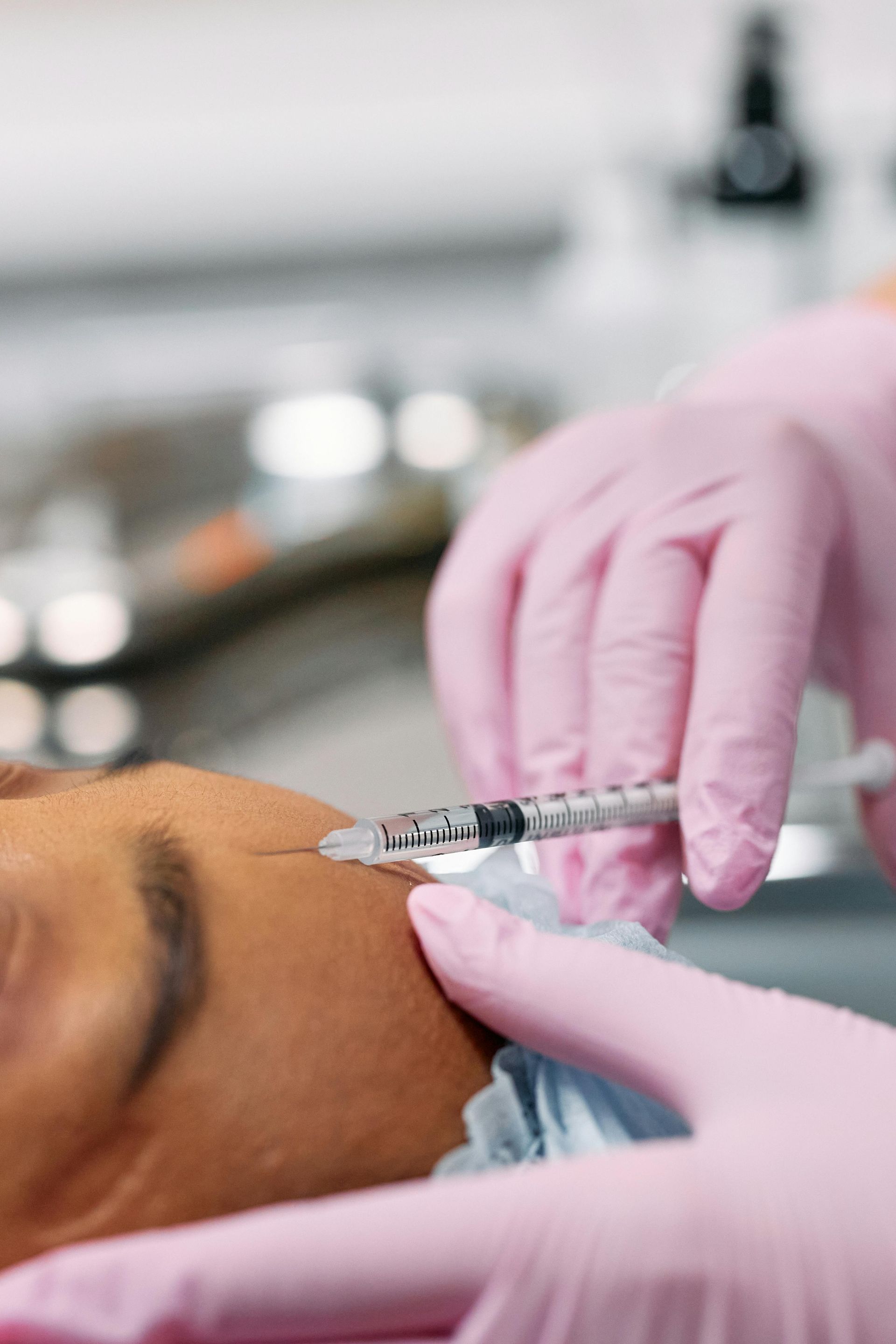 Person receiving a facial injection. Pink-gloved hands hold syringe near face. Clinic setting.