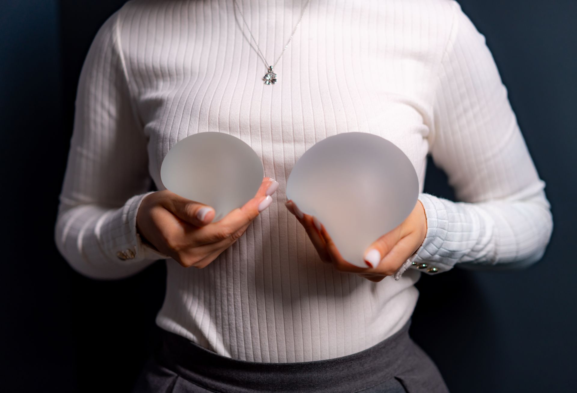 Woman holding two breast implants against her chest, wearing a white sweater.