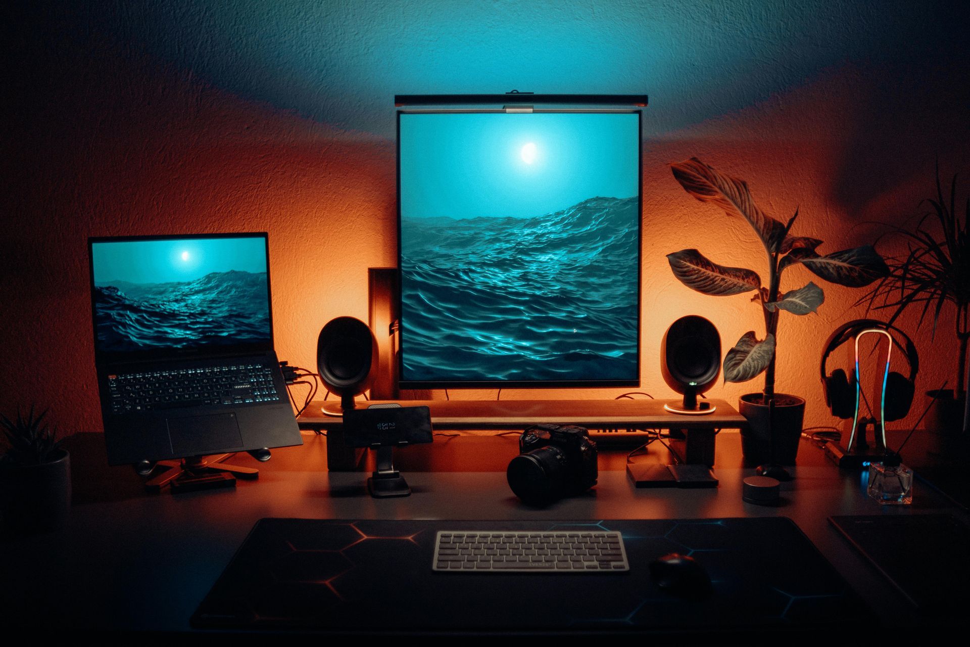 Workspace lit with orange and blue hues. Laptop and large monitor display a landscape. Speakers, camera, and plant accent the desk.