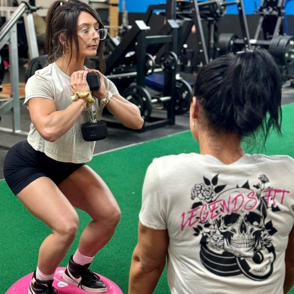 Two women squatting in a gym with one wearing a legends fit shirt