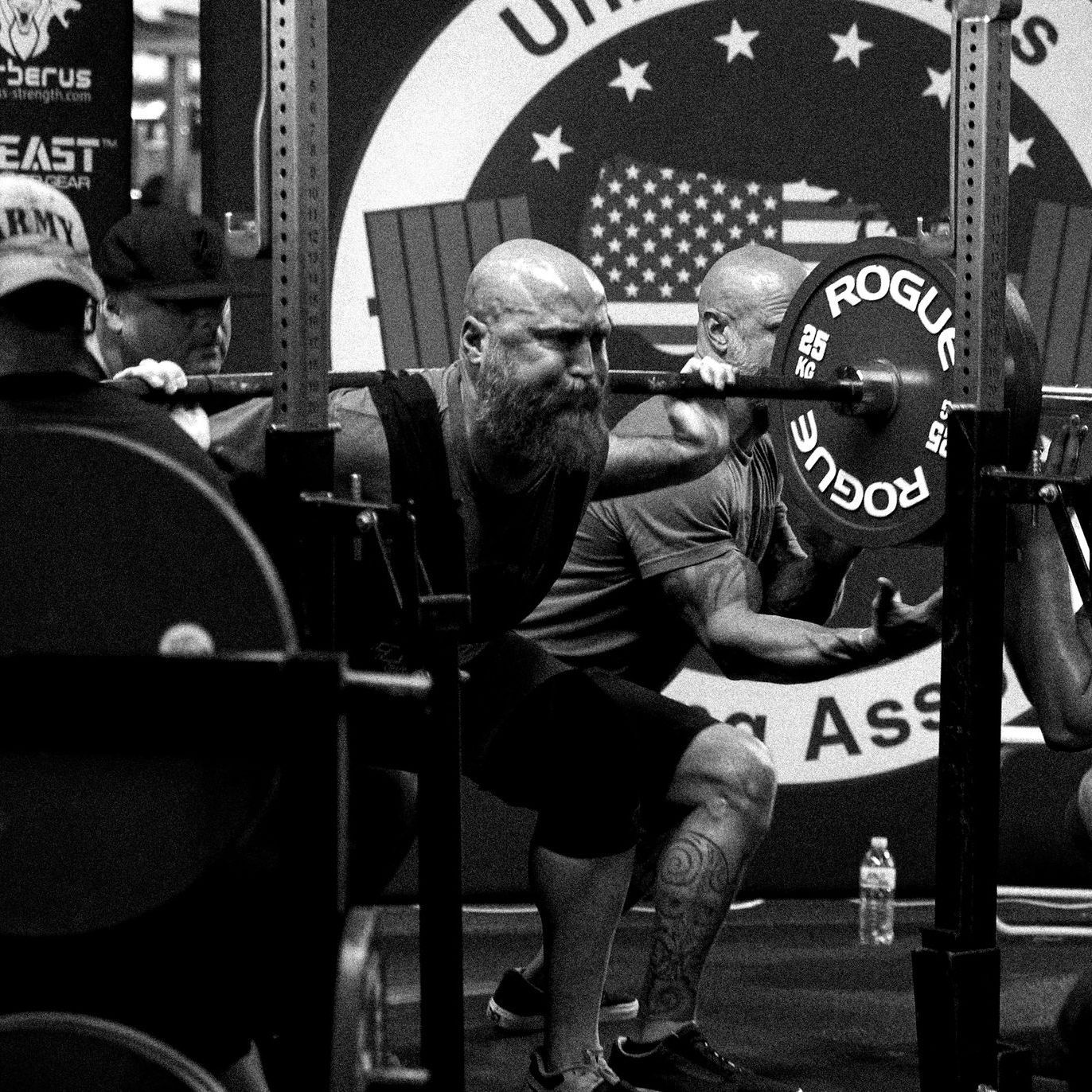 A black and white photo of a man squatting with a rogue barbell.