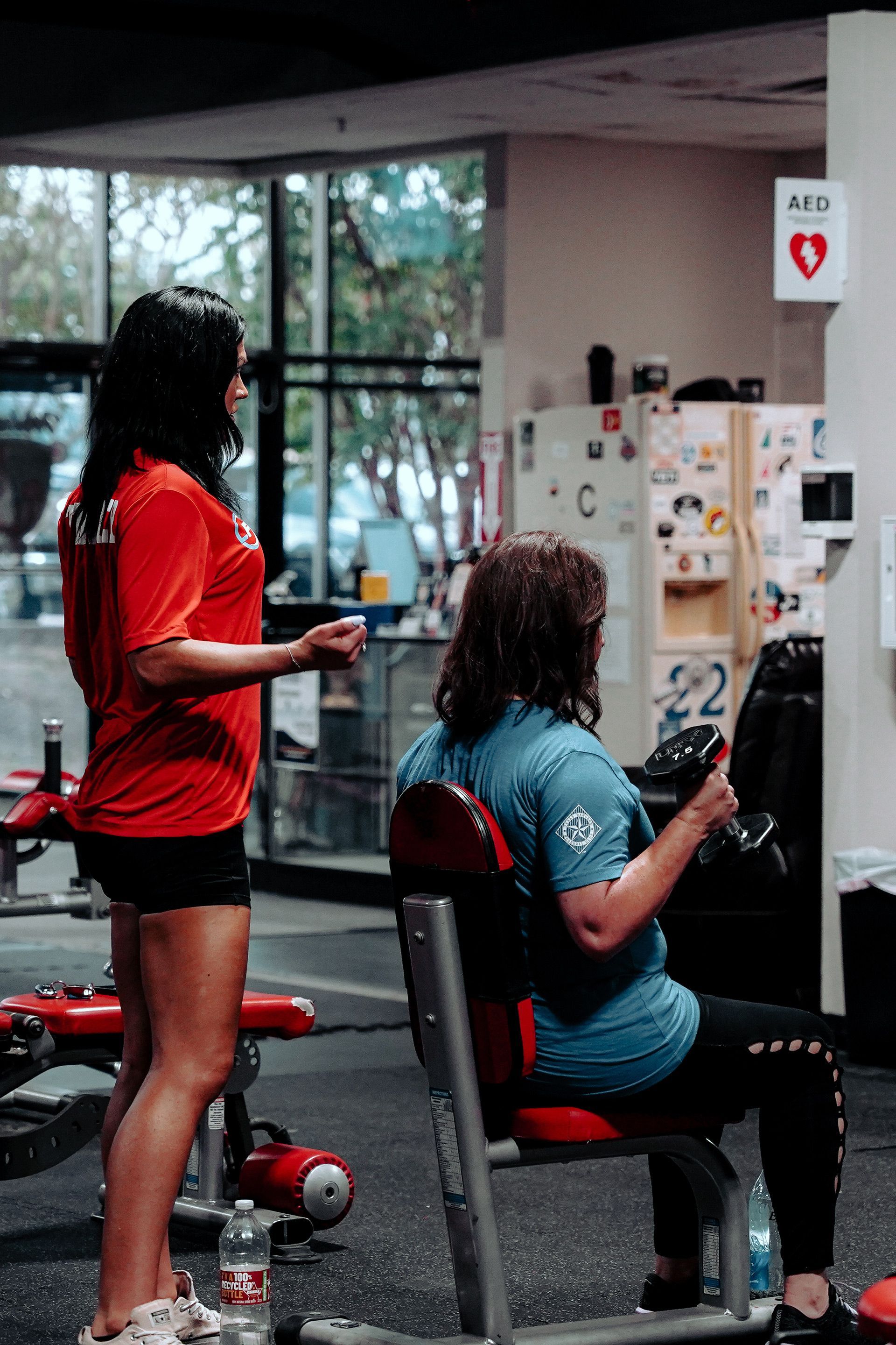 A woman is standing next to a man sitting on a bench in a gym.
