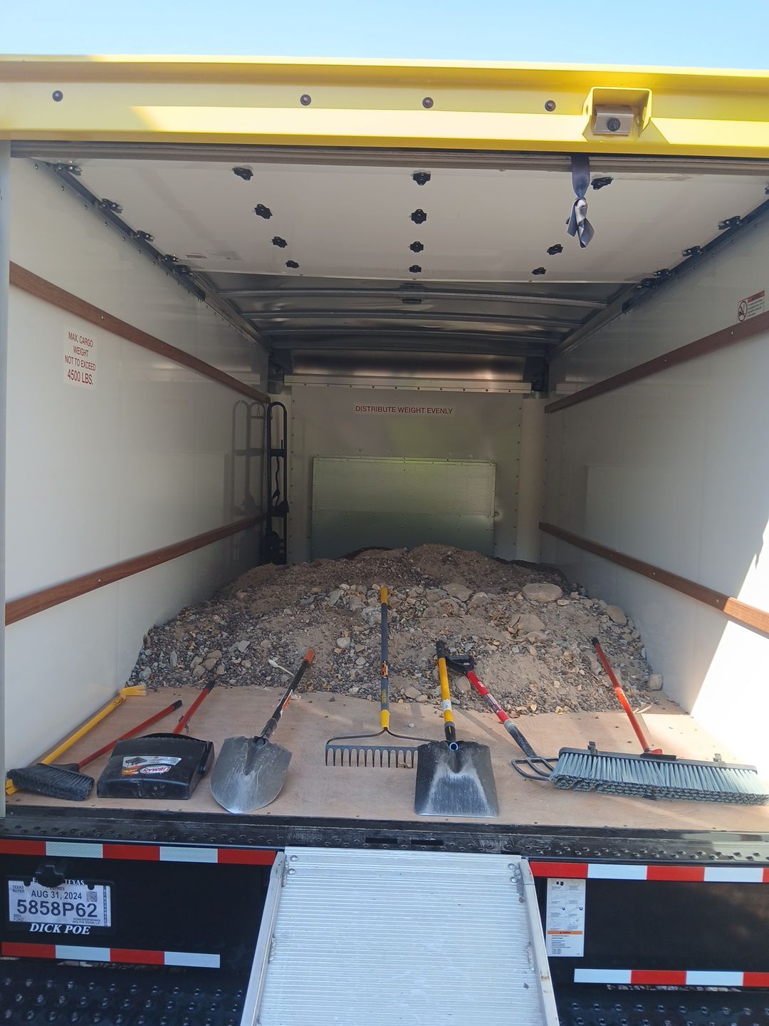 Inside of a truck filled with gravel and tools including shovels, a rake, and a broom.