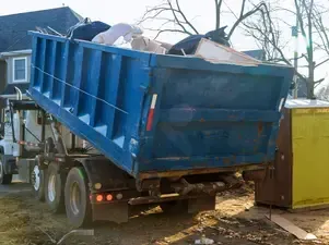 Blue dumpster being lifted by a truck, overflowing with debris, near a house.