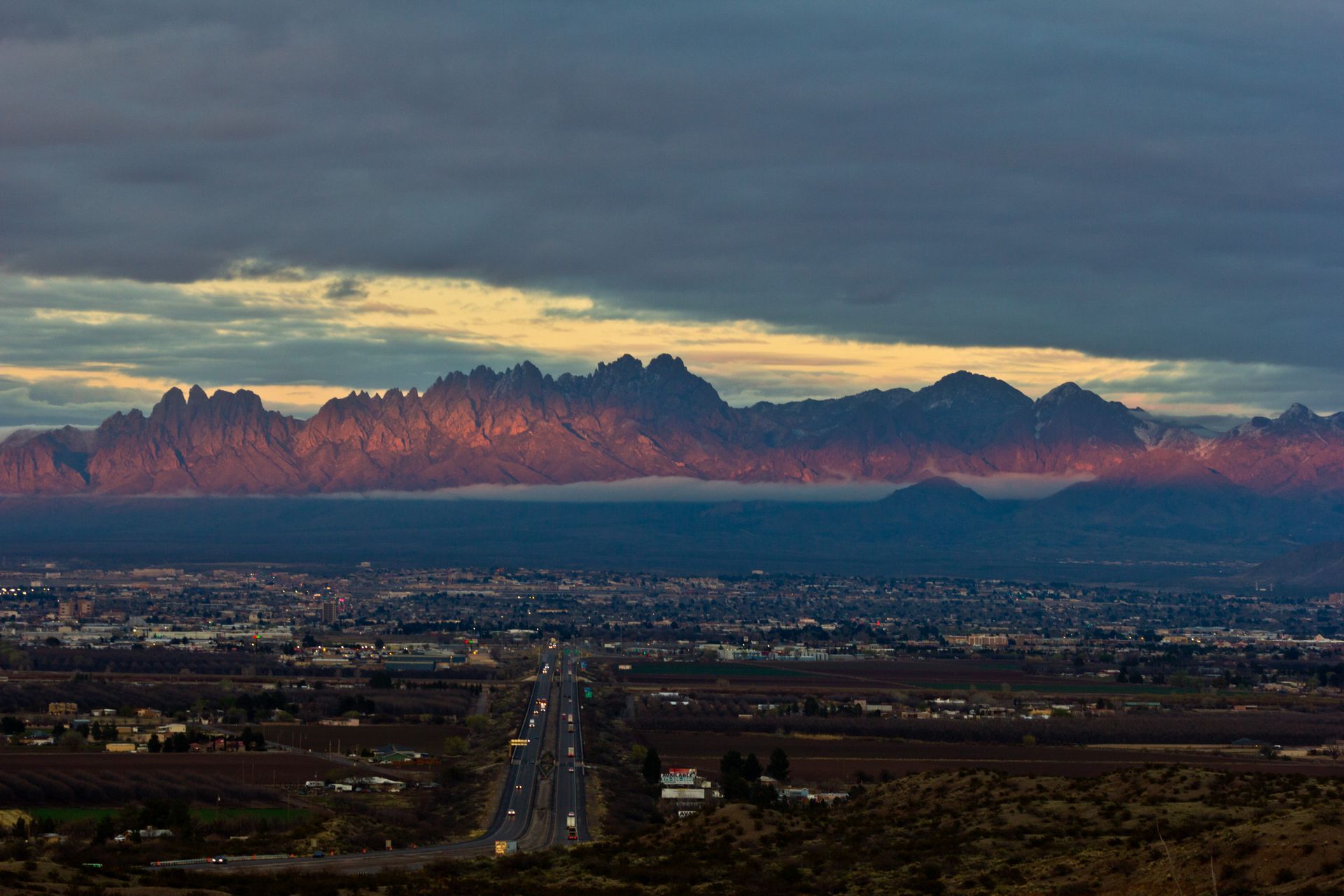 Mountains lit by sunset glow above a city with a road in the foreground under a cloudy sky.