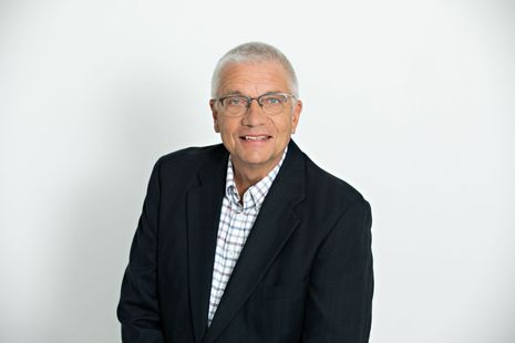 Man with glasses, arms crossed, wearing a maroon shirt with a logo, smiling, neutral background.