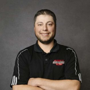Man in black polo shirt with crossed arms smiling. Black background.