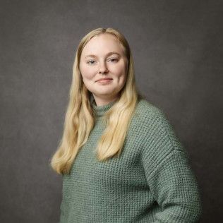 Woman with long blonde hair wearing a green sweater, smiling, against a gray background.