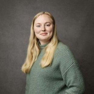 Woman with long blonde hair wearing a green sweater, smiling, against a gray background.