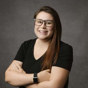 Woman with glasses and black shirt smiles with arms crossed in front of a gray backdrop.