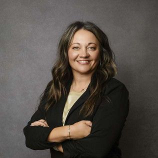 Woman with long brown hair, wearing a black blazer, smiling with arms crossed against a gray background.