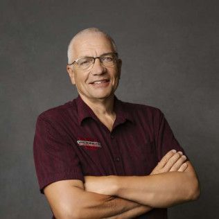 Man with glasses, arms crossed, wearing a maroon shirt with a logo, smiling, neutral background.