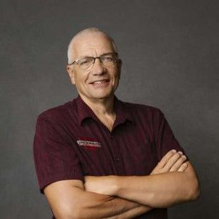 Man with glasses, arms crossed, wearing a maroon shirt with a logo, smiling, neutral background.