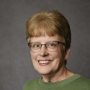 Woman with short, light brown hair and glasses smiles. She wears a green shirt against a gray background.