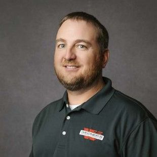 Man with a beard in a dark green polo shirt smiles at the camera. A logo is on the shirt. Gray background.