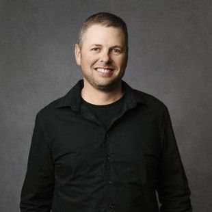 Man smiling, wearing a black shirt, posing against a gray backdrop.