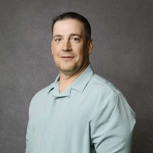 Man in light blue shirt, smiling, against a gray background.
