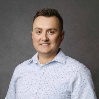 Man with short brown hair smiles, wearing a light blue button-down shirt, against a gray background.