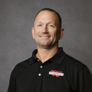 Man with short hair smiles in a black polo shirt with a logo, against a gray background.