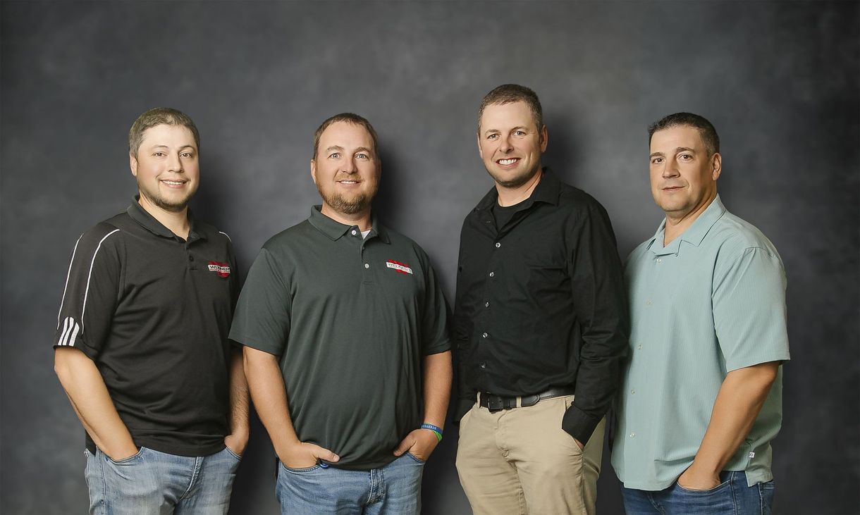 Four men standing in front of a dark gray background. All are wearing shirts and pants, smiling, with hands in pockets.