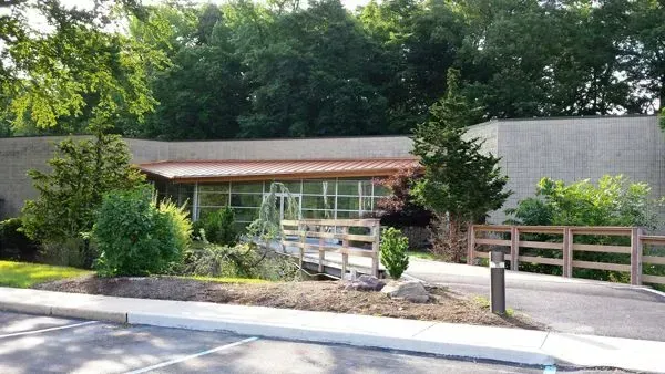 Building with a ramp, greenery, and a wooden fence. Trees in the background, bright sunny day.