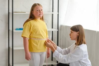 A girl in yellow shirt has waist measured by a woman in white coat indoors.