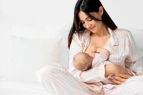 Woman in pajamas breastfeeding baby, smiling. White bedroom setting.