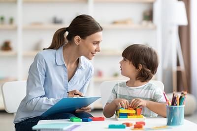 Woman in a light blue shirt smiling at a young boy, who is playing with colorful blocks at a table indoors.
