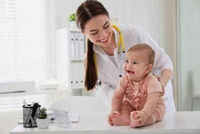 Doctor examines a smiling baby sitting on a white table in a bright office.