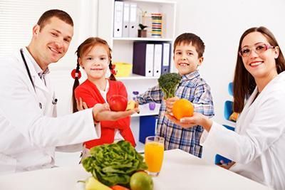 Two doctors with two children, holding fruit and vegetables, smiling in a bright office.