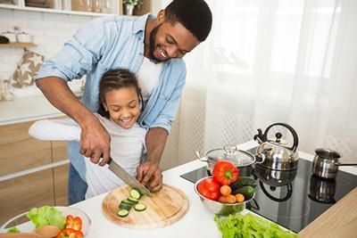 Father and daughter cook in the kitchen. They cut vegetables with a knife, smiling near stove and bowl of ingredients.