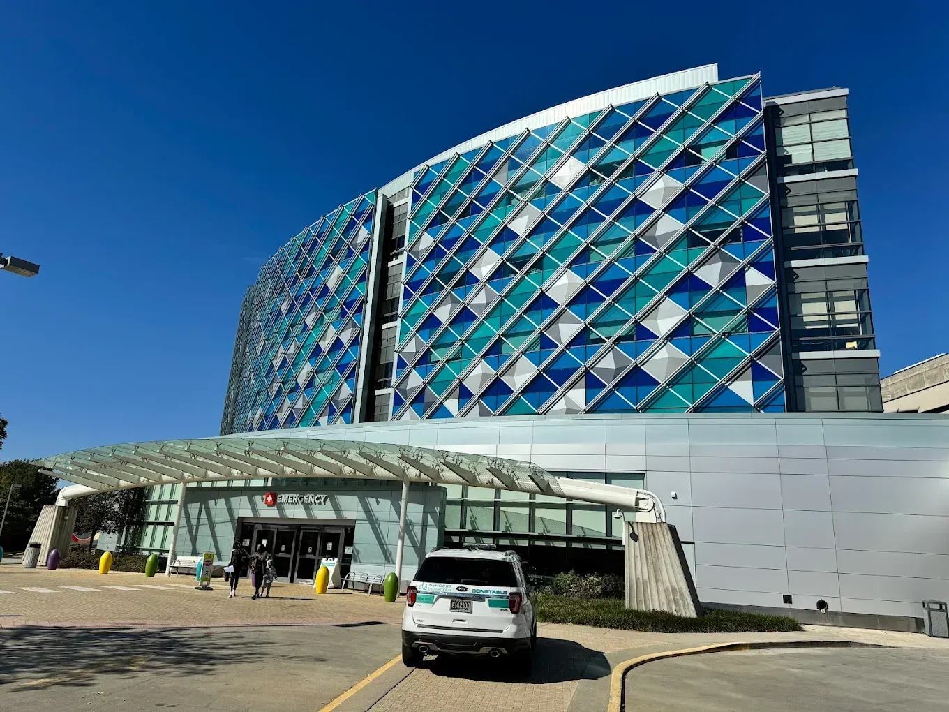 Modern hospital entrance with blue and white patterned facade, people entering, white car in front.
