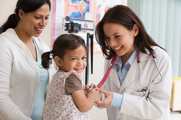 Doctor examining a young girl with a stethoscope, mother watches in a medical office.