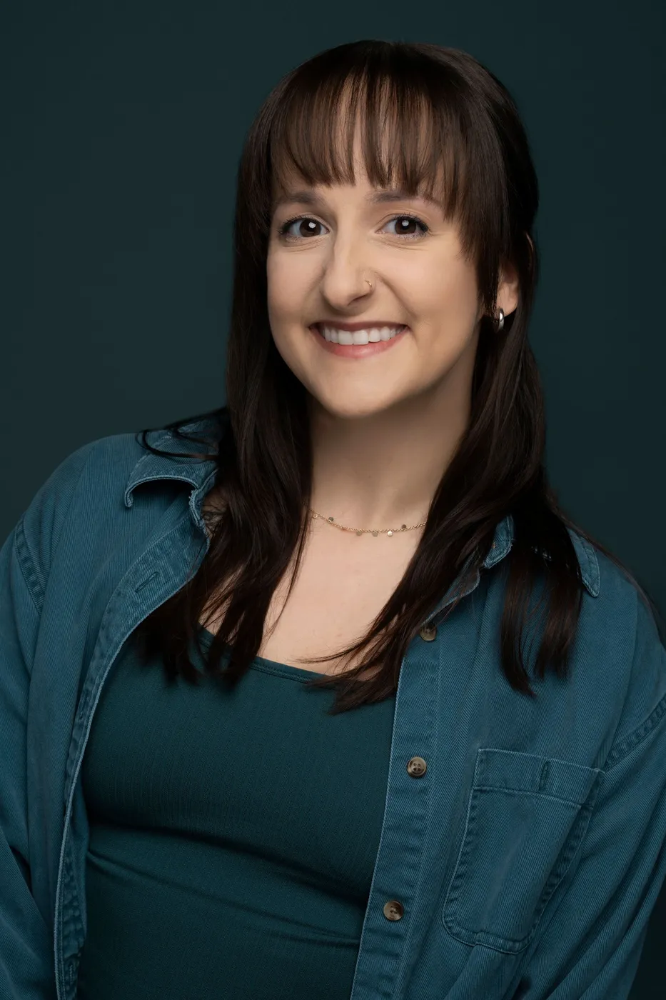 Woman with dark hair and bangs smiles, wearing teal top and denim shirt.