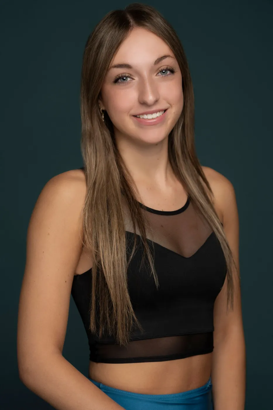 Woman with long brown hair, smiling, wearing a black crop top. Blue background.