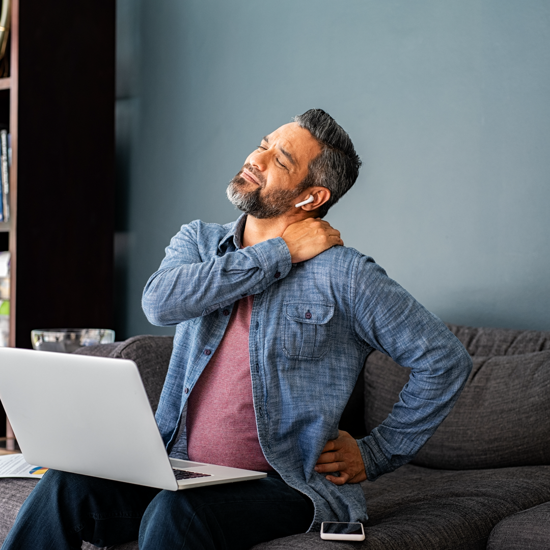 A man is sitting on a couch with a laptop and holding his neck.