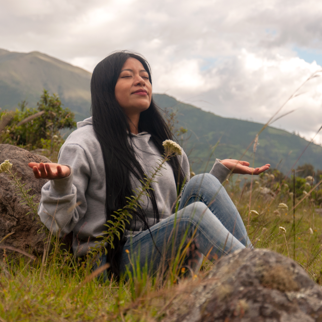 A woman is sitting on a rock in a field with her eyes closed.