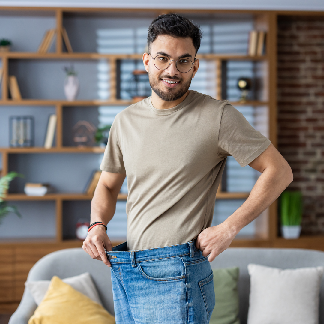 A man is standing in a living room wearing a pair of oversized jeans.