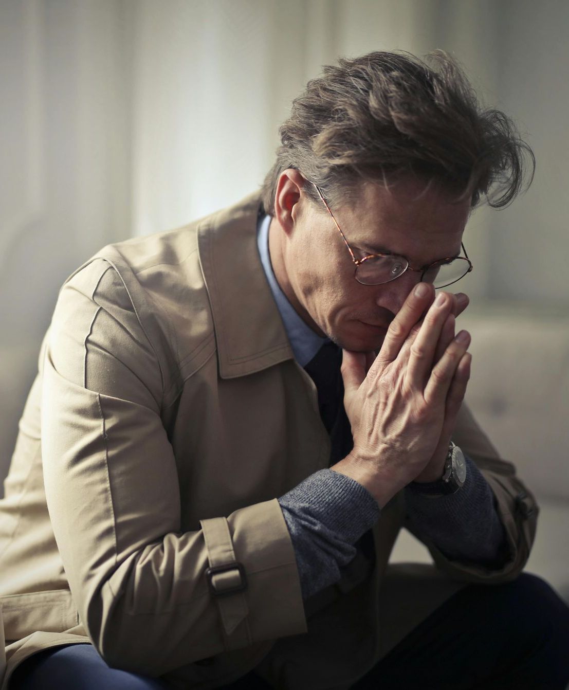A man wearing glasses is sitting on a couch with his head in his hands.