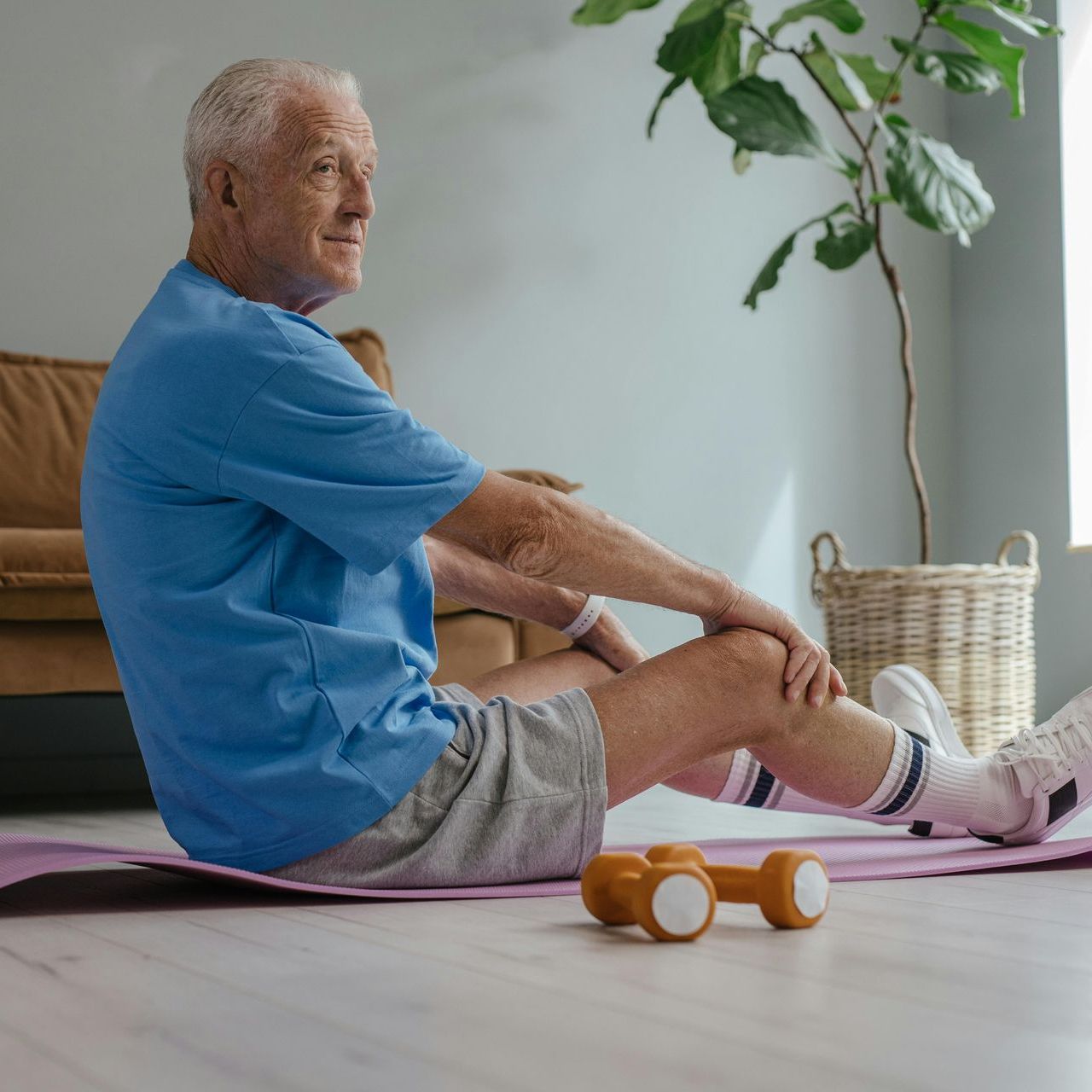 An older man is sitting on a yoga mat next to a pair of dumbbells.